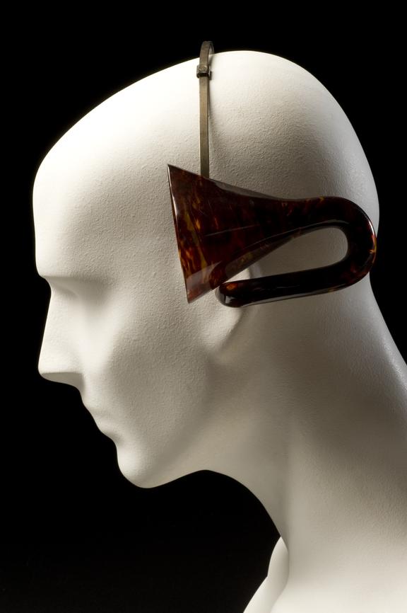 Ear cornets, 2 pairs, for constant wear, imitation tortoiseshell with adjustable head band, early 20th century. Image of ear cornet on a dummy, showing positioning whilst wearing. Black background.